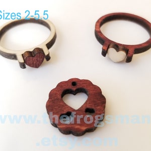 Könnte beinhalten: Zwei Holzringe mit herzförmigen Mittelstücken und ein runder Holzanhänger mit herzförmigem Ausschnitt. Die Ringe zeigen kontrastierende helle und dunkle Holztöne. Der Text auf dem Bild lautet Sizes 2-5.5 und www.thefrogsmane.etsy.com.