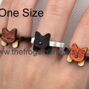 Könnte beinhalten: Drei Holzringe mit Katzenköpfen. Die Ringe sind in Braun, Schwarz und Orange. Der Text "One Size" ist auf dem Bild zu sehen. Der Text "www.thefrogsmane.etsy.com" ist ebenfalls auf dem Bild zu sehen.