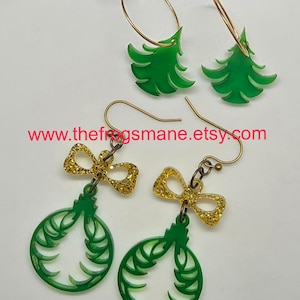 May include: Two pairs of Christmas earrings. One pair features green Christmas tree shapes on gold hoops. The other pair has green ornament shapes with gold glitter bow accents. The text "www.thefrogsmane.etsy.com" is visible.