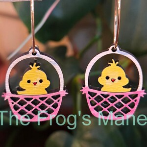 May include: A pair of pink and white wooden earrings with a yellow chick in a basket. The earrings are shaped like a hoop and have a small chick inside a basket.