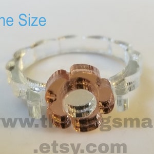 May include: A clear ring with a flower-shaped rose gold centre. The band is clear and textured. The text "One Size" is in the upper left corner. A website address is visible at the bottom.