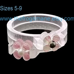 May include: Clear plastic ring with two pink flowers. The flowers have a pearl and a black bead in the center. The ring is available in sizes 5-9. www.thefrogsmane.etsy.com