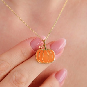 May include: A gold-colored necklace featuring a pumpkin pendant. The pumpkin is orange with a gold outline and stem. The necklace is delicate and appears to be worn around the neck.