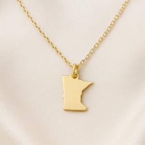 May include: A gold-coloured necklace with a pendant shaped like the state of Minnesota. The pendant hangs from a fine chain. The necklace is shown against a soft, off-white backdrop.