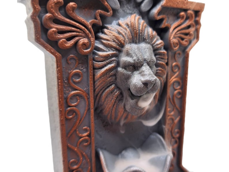 May include: A gray and copper colored incense burner with a lion's head design. The lion's head is carved in detail and has a small opening for incense to be placed. The incense burner is decorated with ornate copper colored designs.