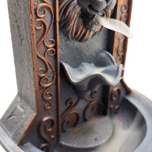 May include: A gray and copper colored incense burner shaped like a lion's head with intricate details. The incense burner has a small bowl for incense and a larger bowl below to catch the ash. Smoke is rising from the lion's mouth.