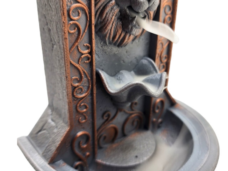May include: A gray and copper colored incense burner shaped like a lion's head with intricate details. The incense burner has a small bowl for incense and a larger bowl below to catch the ash. Smoke is rising from the lion's mouth.