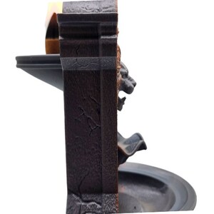 May include: A black and brown tabletop fountain with a lion head design. The fountain has a tiered design with a small basin at the top and a larger basin at the bottom. The fountain is made of a textured material that resembles stone.