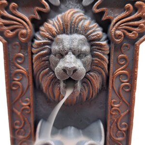 May include: A gray and copper colored incense burner with a lion's head design. The incense burner is shaped like a rectangular frame with ornate details. Smoke is coming out of the lion's mouth.