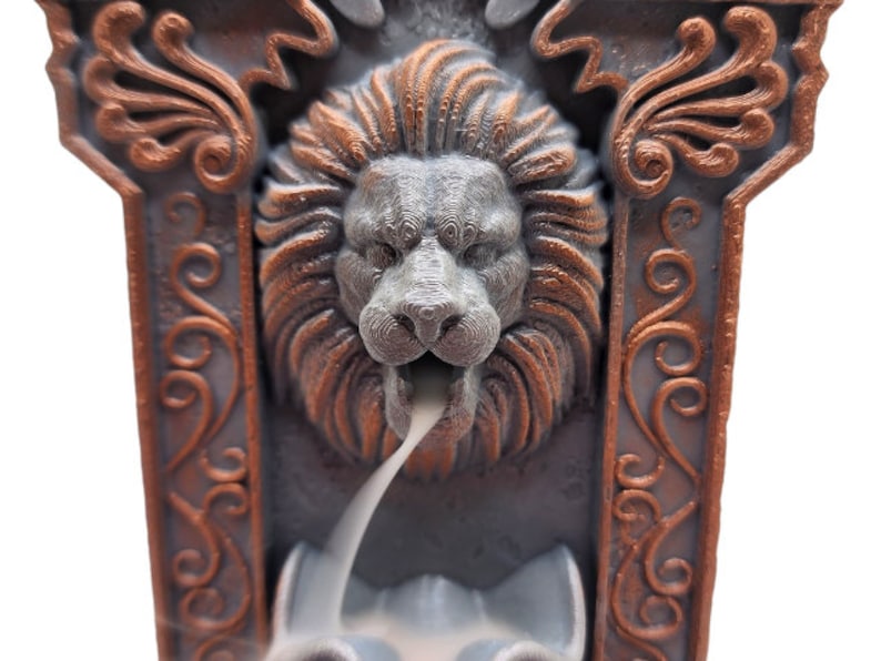May include: A gray and copper colored incense burner with a lion's head design. The incense burner is shaped like a rectangular frame with ornate details. Smoke is coming out of the lion's mouth.
