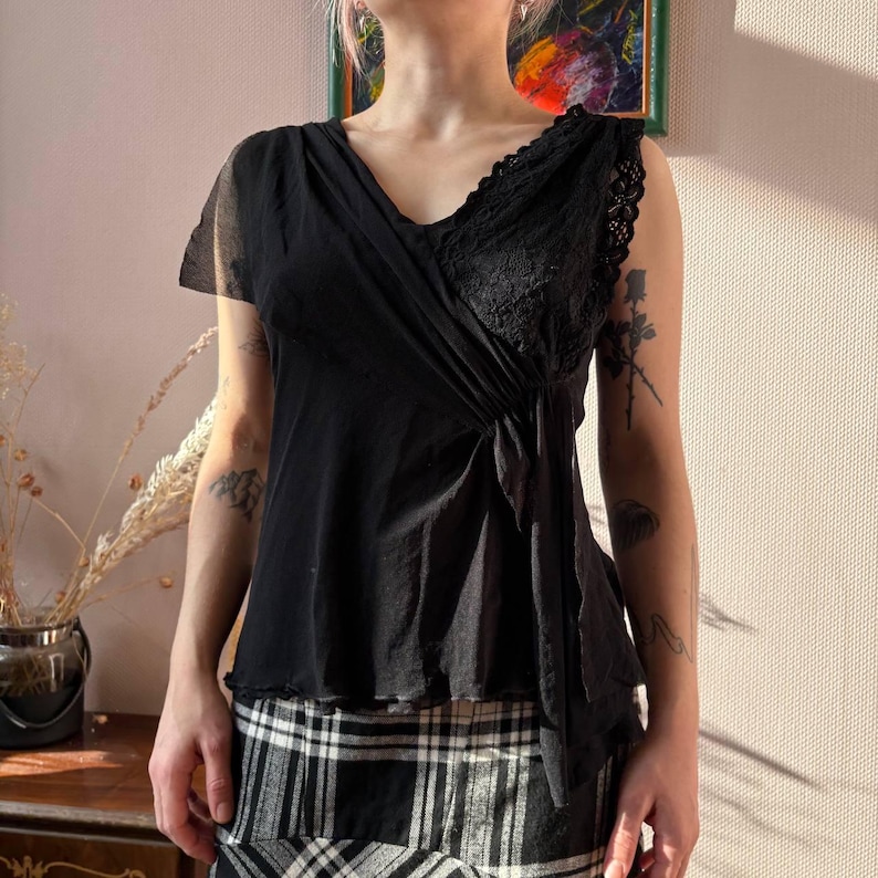 May include: Black sleeveless top with a V-neckline and lace detailing on the right shoulder. The top has a draped design across the front and a slightly ruffled hem. The model is wearing a black and white plaid skirt.