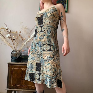 May include: A patterned midi dress with a halter neckline. The dress features a beige background with black and teal geometric designs. The dress has a fitted bodice and a slightly flared skirt, with a ruffled hemline. The dress is made of a lightweight fabric.