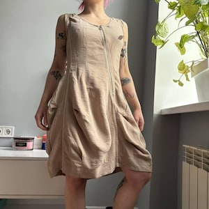 May include: A beige sleeveless dress with a front zipper closure. The dress has a unique, draped design with gathered fabric details. The dress is made of a lightweight material, perfect for warm weather.