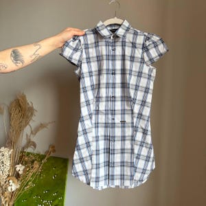 May include: A short-sleeved plaid shirt in white, blue, and gray, hanging on a white hanger. The shirt has a button-down front and a small logo near the hem. The sleeves have a slight puff detail.