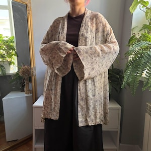 May include: A lightweight, open-front kimono-style cardigan in a cream colour featuring a delicate, dark blue floral line-art pattern. The garment has long, wide sleeves and a loose, flowing fit that drapes over a dark brown top and black trousers.