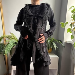 May include: A black long-sleeved cardigan featuring a textured knit pattern, a hooded collar, and frayed trim along the front edges. The garment includes a tie closure at the neckline and a layered, ruffled hemline that extends past the waist.