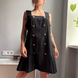May include: A sleeveless black mini dress featuring a square neckline and a double-breasted front design with two vertical rows of metallic gold buttons. The garment has a pleated hemline that adds volume to the lower portion of the dress.