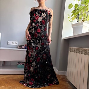 May include: A floor-length black dress featuring a vibrant floral pattern in shades of red, white, and grey. The garment has thin spaghetti straps, a ruffled neckline detail, and a straight silhouette that extends to the floor with a hem.