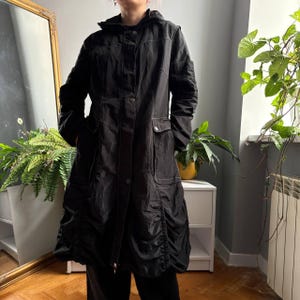 May include: A black long-length hooded jacket featuring a front button closure, long sleeves, and two large front pockets. The garment includes visible white contrast stitching and gathered, ruched detailing along the lower hemline.
