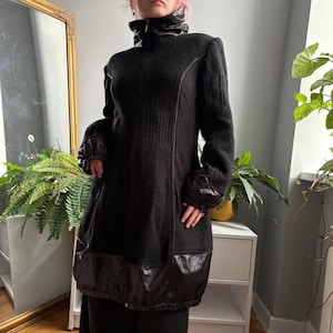 May include: A black long-sleeve coat featuring a high collar, a full-length front zipper, and a textured, shiny panel along the bottom hem. The garment includes matching shiny fabric accents on the sleeve cuffs and vertical seam detailing.