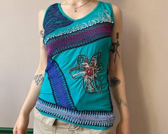 May include: A turquoise sleeveless top with a V-neck and a floral design. The top features a patchwork design with blue, purple, and white embroidered details. The bottom hem and neckline are trimmed with a decorative stitch.