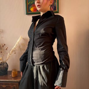 Puede incluir: Una camisa negra abotonada con mangas largas y cuello clásico. La camisa tiene una silueta entallada y puños anchos. La prenda está hecha de una tela oscura y lisa, y forma parte de una colección de ropa.