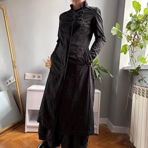 May include: A long-sleeved black coat featuring a high collar, a front button closure, and side pockets. The garment has a fitted bodice that transitions into a flared, floor-length skirt, constructed from a solid black fabric.