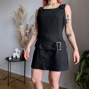 May include: A dark gray sleeveless dress with a square neckline and a belt detail. The dress features a buckle closure and a fitted silhouette. The fabric appears to be a textured material. The dress is a mini length.