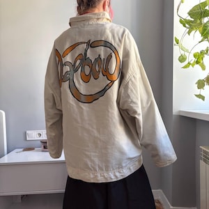 以下が含まれることがあります： ベージュの襟付き長袖ジャケット。 ジャケットの背面には、オレンジとグレーのデザインで「Barbour」の文字が描かれています。 ジャケットはニュートラルカラーで、軽量素材で作られているようです。