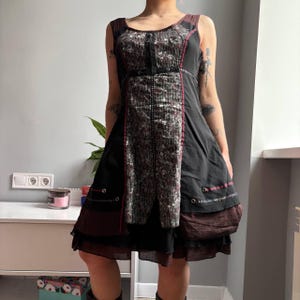 Puede incluir: Vestido negro sin mangas con diseño patchwork. El vestido presenta un panel central estampado, detalles burdeos y una falda en capas. Los detalles incluyen costuras decorativas y un cinturón pequeño.