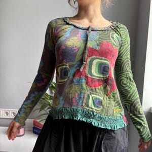 May include: A long-sleeved top with a unique floral design. The top features a mix of colors, including red, green, blue, and purple, with a ruffled neckline and hem. The right sleeve has a green swirl pattern.