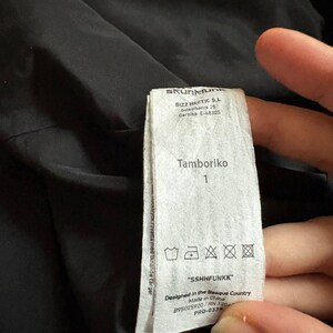 K&ouml;nnte beinhalten: Ein wei&szlig;es Kleidungsetikett mit dem Text "Tamboriko" und der Zahl "1". Das Etikett enth&auml;lt auch Waschsymbole und den Text "SSHHFUNKK". Das Etikett befindet sich vor einem schwarzen Hintergrund.
