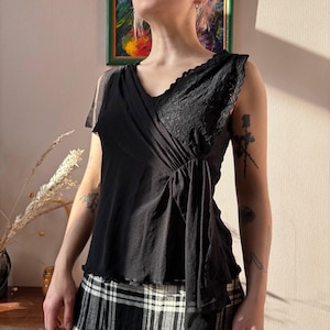 May include: Black sleeveless top with a V-neckline and lace detailing on the right shoulder. The top has a draped design and a ruffled hem. The model is wearing a black and white plaid skirt.