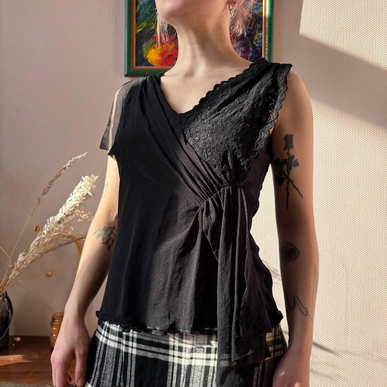 May include: Black sleeveless top with a V-neckline and lace detailing on the right shoulder. The top has a draped design and a ruffled hem. The model is wearing a black and white plaid skirt.