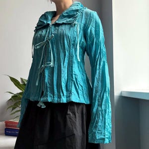 May include: A turquoise long-sleeved jacket with a ruffled collar and tie fastenings. The jacket features a textured, pleated design and is worn with a black skirt. The sleeves are long and gathered at the wrists.