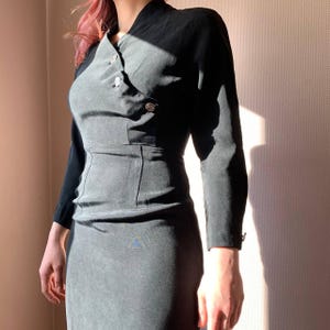 May include: A gray and black dress with a long sleeve and a button closure. The dress has a fitted silhouette and a high neckline.