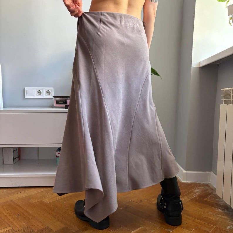 May include: A long, light gray skirt with a smooth, matte texture. The skirt features a high-waisted design with vertical seam detailing that creates a flared, asymmetrical hemline, providing a flowing silhouette that reaches below the knees.