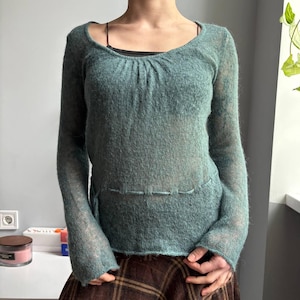 Op de afbeelding: Een tealkleurige trui met lange mouwen, een ronde hals en een geplooid detail bij de halslijn. De trui heeft een semi-transparante, getextureerde uitstraling en een gestikt detail in de taille. De mouwen zijn lang en licht uitlopend.
