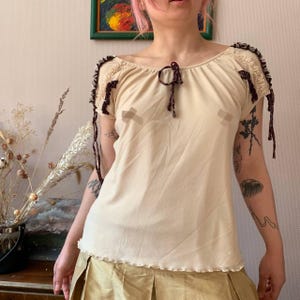 Pode incluir: Uma blusa creme de manga curta com decote redondo. A blusa apresenta acabamentos em castanho escuro ao longo das mangas e um laço a condizer no decote. A bainha inferior tem uma borda ondulada. A blusa é combinada com uma saia dourada.