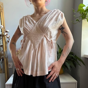 May include: A pale pink sleeveless blouse featuring a V-neckline, a central diamond-shaped panel, and gathered fabric details. The garment includes side ties at the waist and a flared silhouette, constructed with a lightweight, semi-sheer fabric.