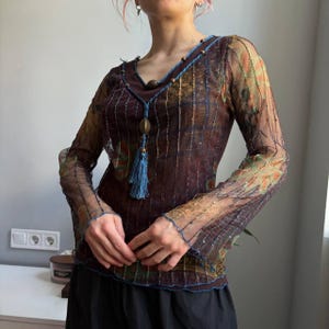 Op de afbeelding: Een doorschijnende top met lange mouwen in donkerbruin met een veelkleurig bloemenpatroon, voorzien van een V-hals met blauwe koordafwerking, houten kralen en een blauw kwastje. Het kledingstuk heeft golvende randen bij de mouwen en de zoom.