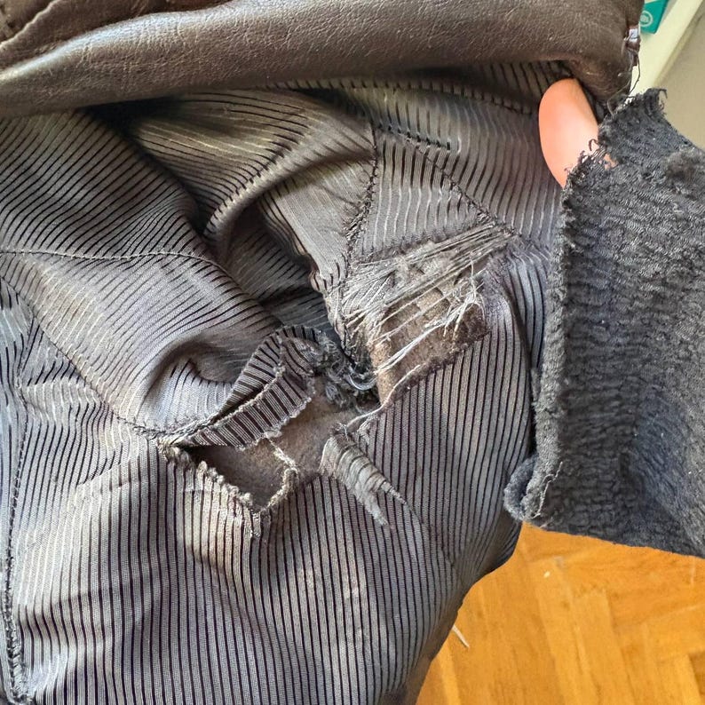 May include: Close-up of the torn lining of a brown leather jacket. The interior fabric is a dark gray with vertical pinstripes. The jacket's leather exterior is visible at the top. The sleeve cuff is visible on the right side.