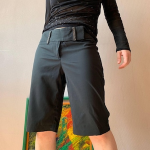 May include: Black Bermuda shorts with a wide waistband adorned with small, sparkling embellishments. The shorts have a tailored fit and reach the knees. The model is wearing a black long-sleeved top.