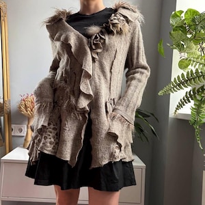 May include: A beige knit cardigan featuring a ruffled collar, long sleeves with frayed edges, and a textured surface. The garment includes a front zip closure, layered fabric panels, and a subtle leopard print pattern on the lower side section.