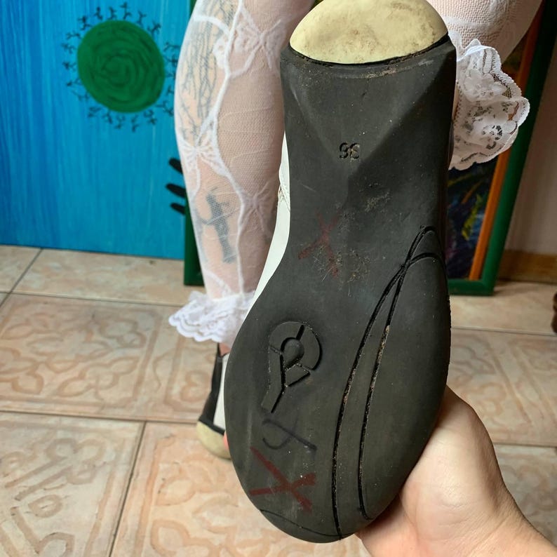 Puede incluir: Primer plano de la suela de goma negra de un zapato de cuero blanco. La suela tiene un patr&oacute;n texturizado y una marca roja en "X".