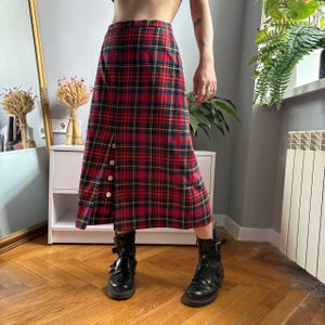 Könnte beinhalten: Ein wadenlanger Rock mit einem rot-grün-blauen Schottenkaro-Muster. Der Rock hat eine hohe Taille und einen vertikalen Schlitz auf der Vorderseite, der mit vier kleinen, runden, hellen Knöpfen entlang der Öffnungskante akzentuiert ist.
