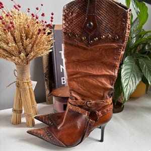 May include: A brown leather knee-high boot with a pointed toe, featuring a textured snakeskin-patterned upper shaft and toe cap. The boot includes decorative metal studs, a wrap-around buckled strap at the ankle, and a slim high heel.