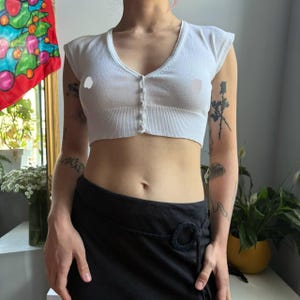May include: A white cropped cardigan featuring short sleeves, a deep V-neckline, and a front button closure with five matching white buttons. The garment has a ribbed hem and a fitted silhouette, crafted from a knit fabric with a smooth texture.