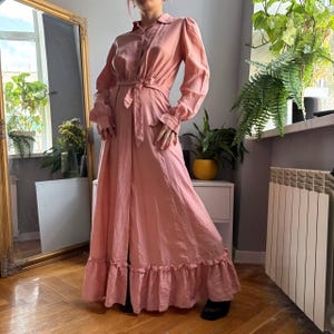 May include: A long-sleeved, floor-length dusty pink dress featuring a button-down front, a collared neckline, and a matching fabric tie belt at the waist. The garment includes gathered cuffs at the wrists and a ruffled hemline at the bottom edge.