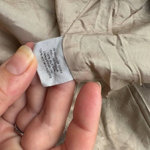 May include: Close-up of a tag with text. The tag reads "RN 66170 CA 32054 SHELL/EXTERIEUR 100% SILK/SOIE LINING/DOUBLURE 100% ACETATE/ACETATE". The tag is white with black text. The background is a tan fabric.